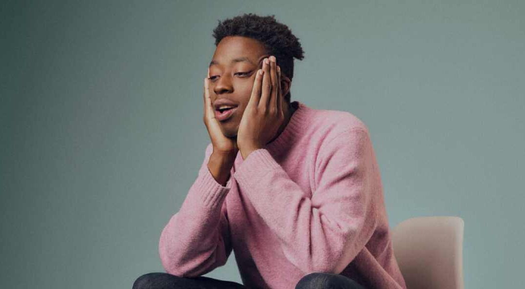 Ayo Adenekan, a Black man, sits on an armchair with his face in his hands, in a studio photoshoot against a blue/grey background